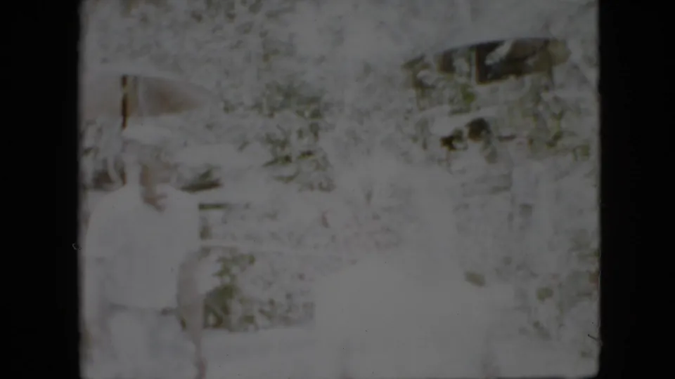 This 1958 Pennsylvania home-movie still appears to show a person in pale clothing near dense foliage and a partial garden structure, with severe highlight fading.