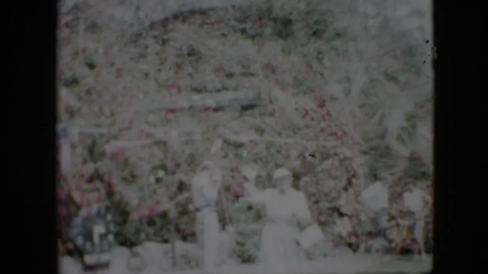 Faded 1958 Pennsylvania 8mm frame with several onlookers faintly visible near dense garden foliage and bright outdoor light.