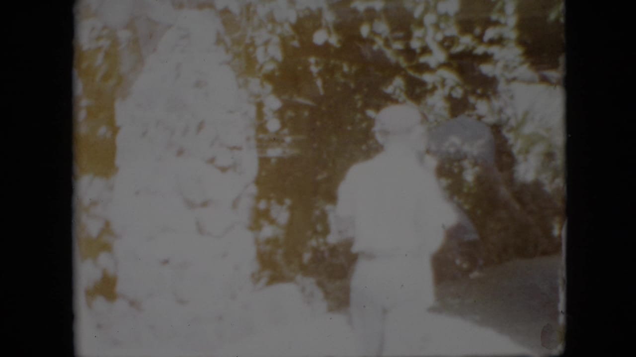 Two standing figures outdoors, including a person in a light dress and a man in a hat.