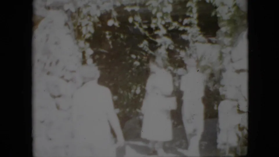 1958 Pennsylvania home-movie frame with three adults near a tree, including a figure in a light dress and a hat-wearing person, rendered with heavy exposure and film grain.