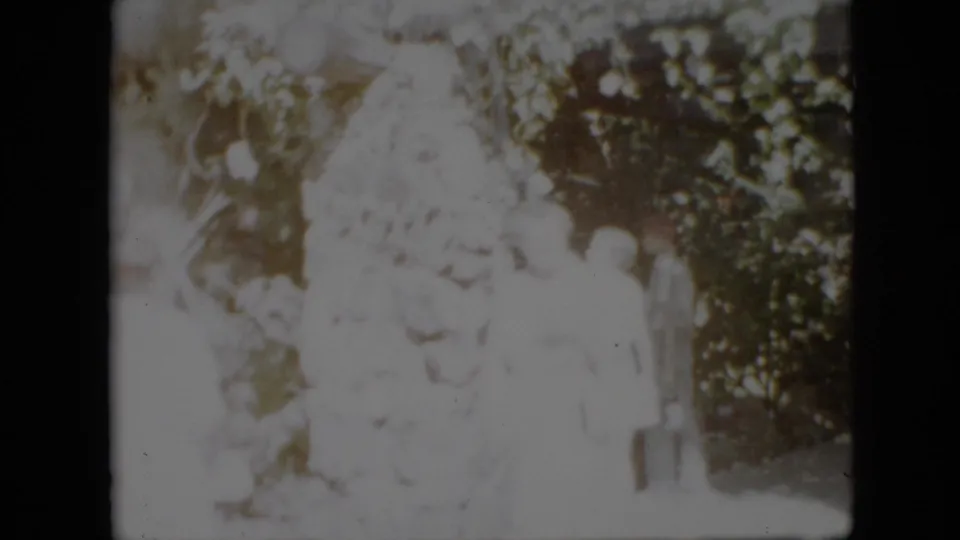 Two women stand close together near the large tree and dense yard foliage, continuing the apparent greeting moment from the 1958 Pennsylvania home movie.