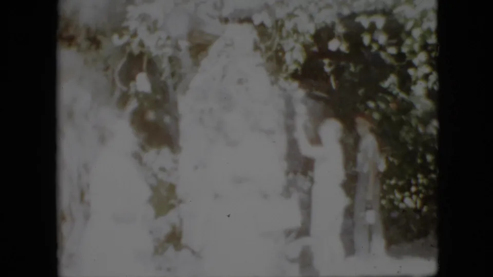 1958 Pennsylvania home-movie frame showing several people gathered in a wooded setting near a large pale statue, with strong overexposure obscuring facial detail.
