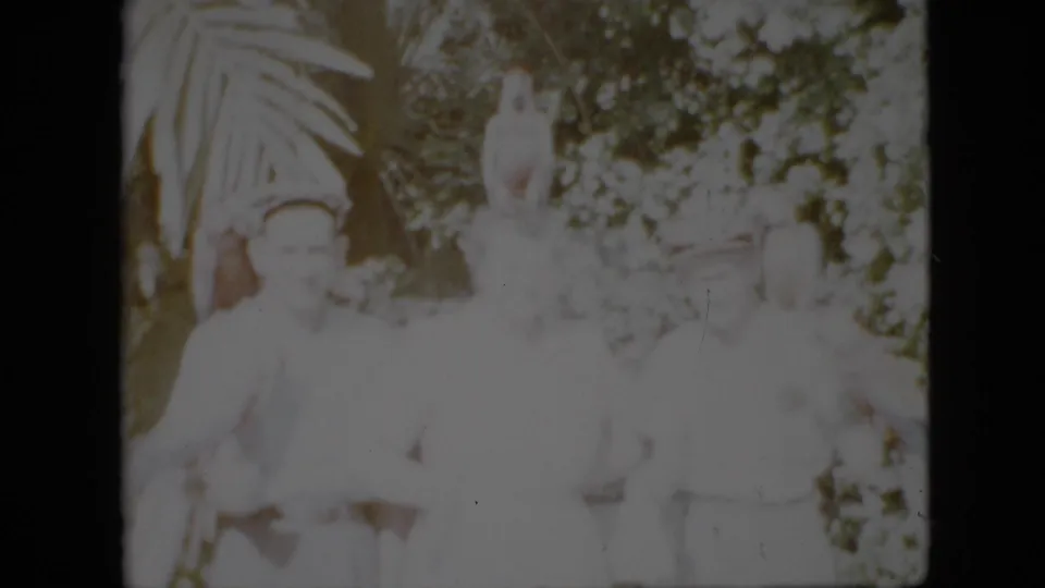 1958 Pennsylvania frame with adults in light clothing gathered near foliage and a tall pale garden statue or sculpture, captured in faded 8mm color.
