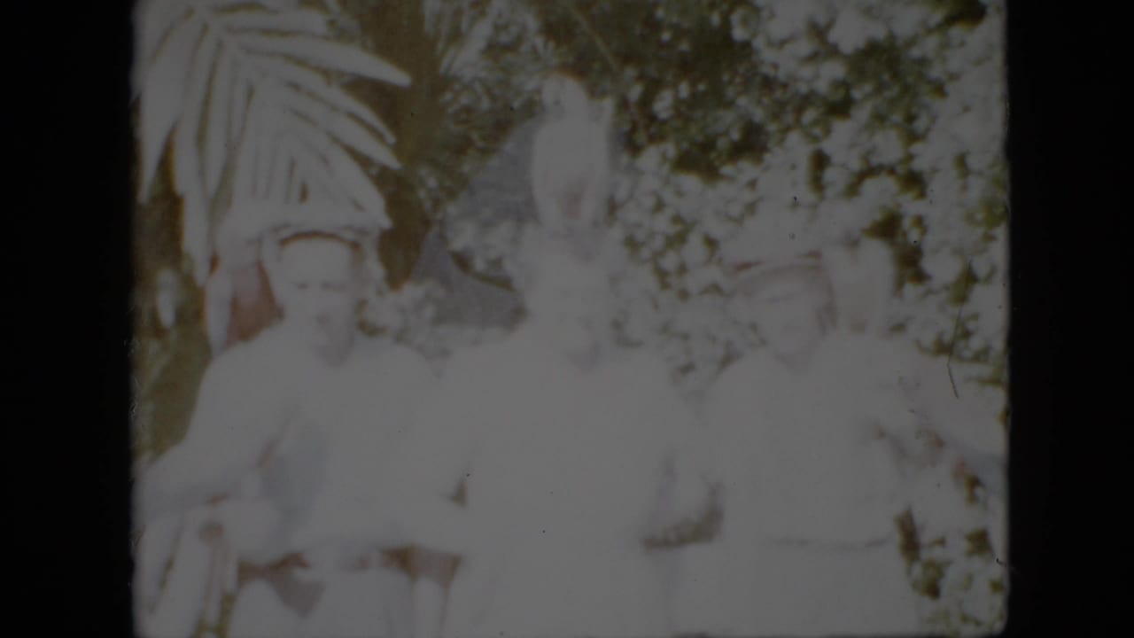 1958 Pennsylvania 8mm home-movie frame showing several adults in pale clothing posed outdoors near parrots, with broad fronds and dense foliage behind them.