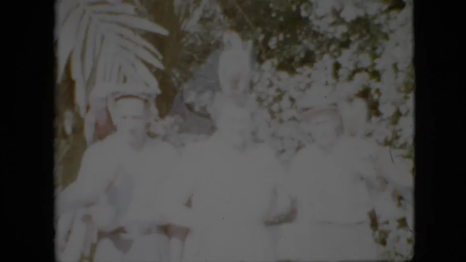 1958 Pennsylvania 8mm home-movie frame showing several adults in pale clothing posed outdoors near parrots, with broad fronds and dense foliage behind them.
