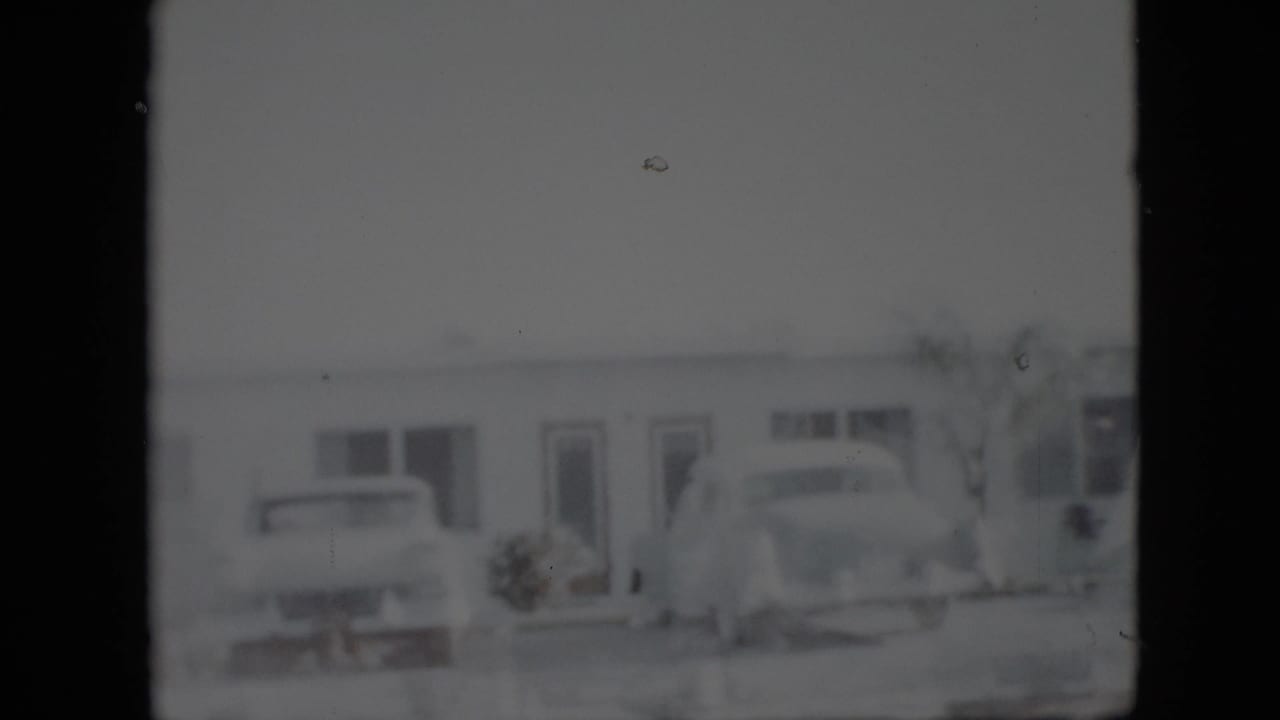 A soft 8mm view of a low Pennsylvania building in 1958 with two parked mid-century cars near the entrance.
