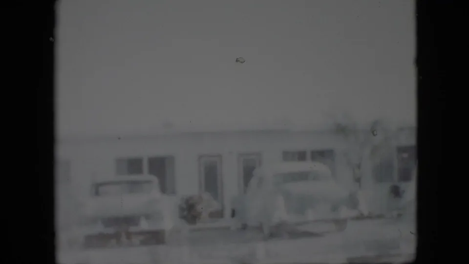 A soft 8mm view of a low Pennsylvania building in 1958 with two parked mid-century cars near the entrance.