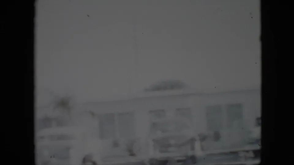Soft black-and-white 1958 Pennsylvania home-movie frame showing parked mid-century cars outside a low building with windows and a doorway.