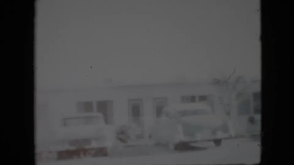 Black-and-white home-movie frame showing a low Pennsylvania building frontage with parked cars and indistinct doors or windows.