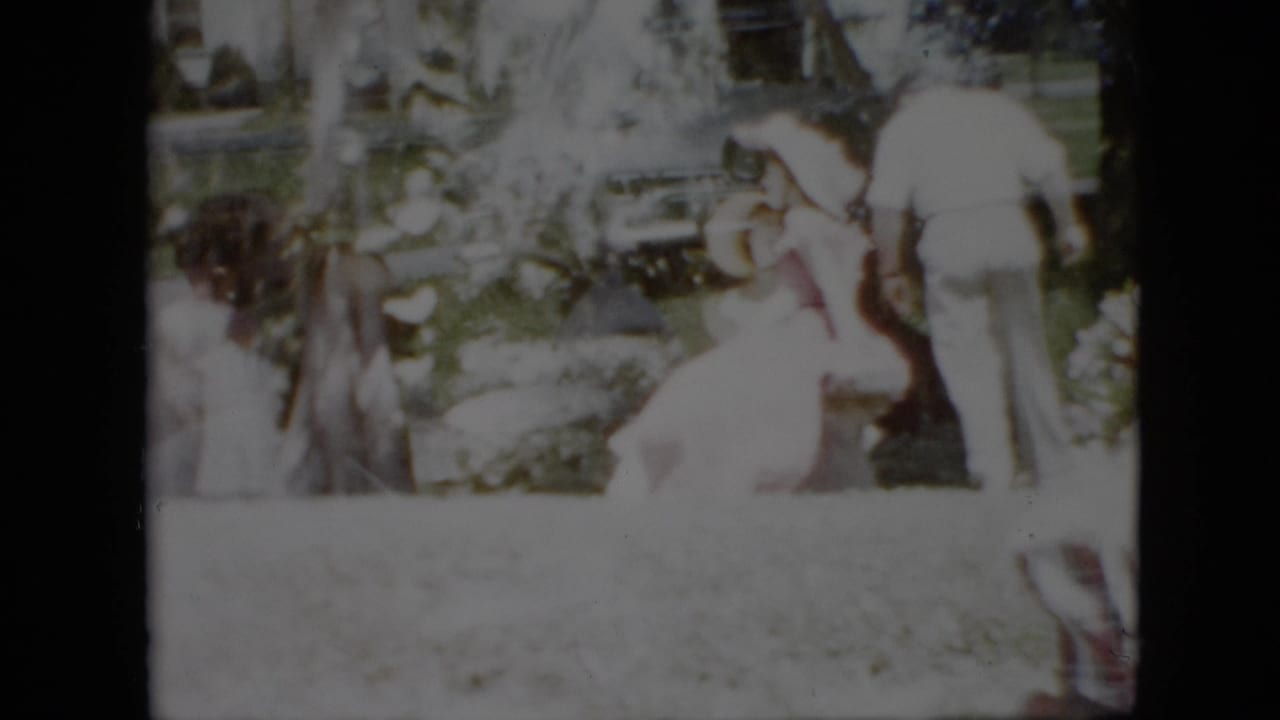 1958 Pennsylvania home-movie frame showing adults in light summer clothing gathered on a green lawn beside garden plantings, with a parked car partly visible behind them.