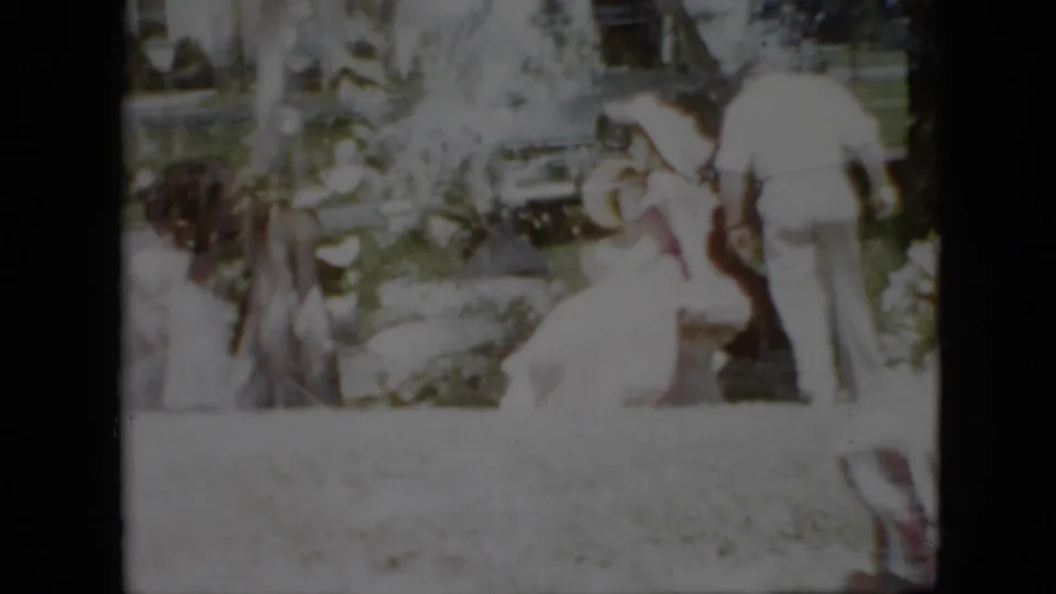 1958 Pennsylvania home-movie frame showing adults in light summer clothing gathered on a green lawn beside garden plantings, with a parked car partly visible behind them.
