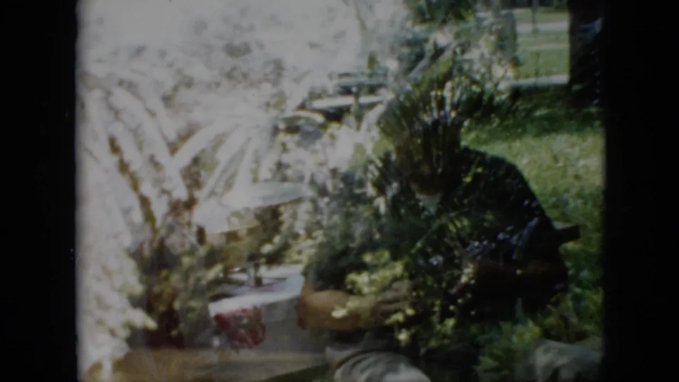 1958 Pennsylvania home-movie frame showing a garden table, potted plants, dense foliage, lawn, and a seated adult partly obscured by leaves and shadow.