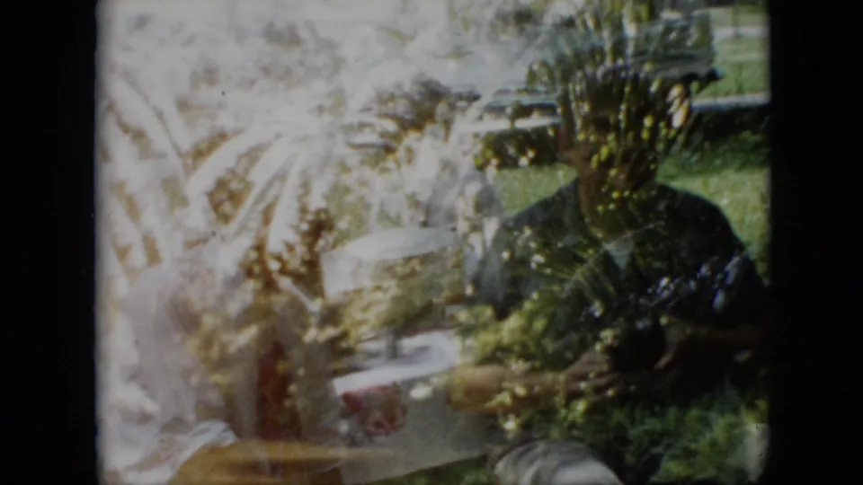 1958 Pennsylvania home-movie frame with a seated adult near potted plants and a garden table, surrounded by bright foliage and green lawn in a backyard setting.