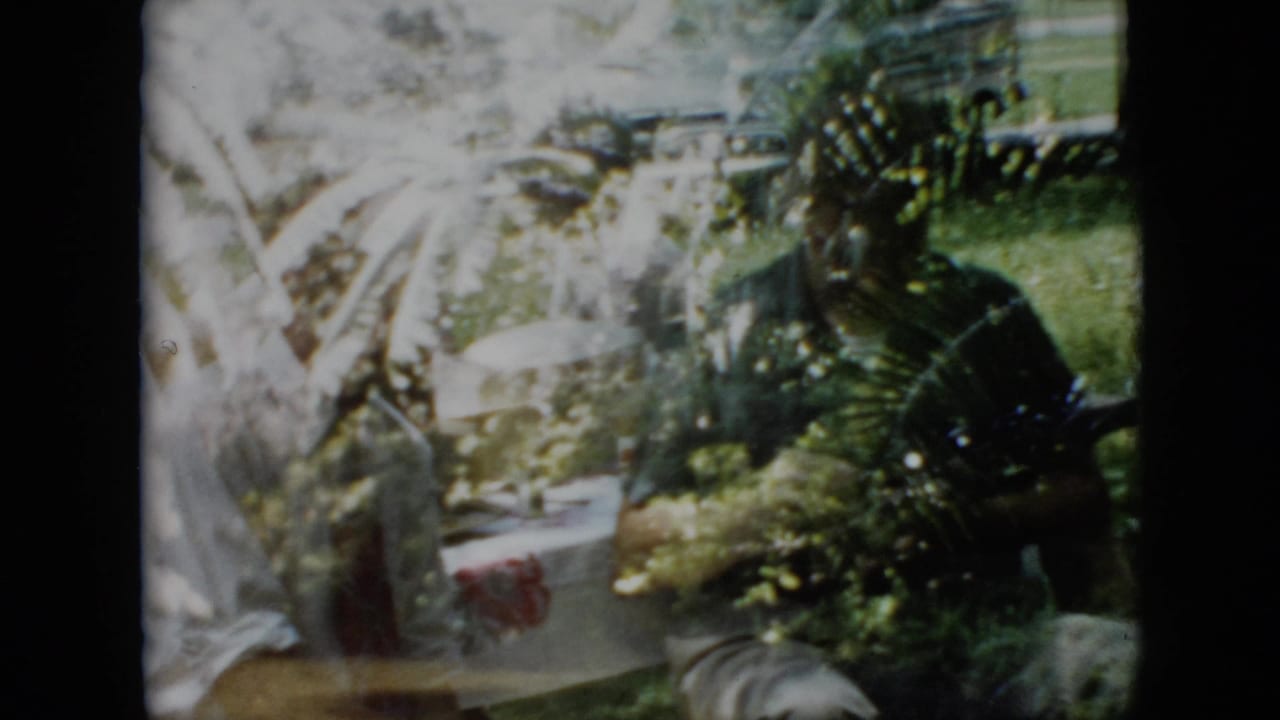 Pennsylvania 1958 home-movie frame showing adults at an outdoor family picnic table, with a white tablecloth, plates, greenery, and a parked car shape in the background.