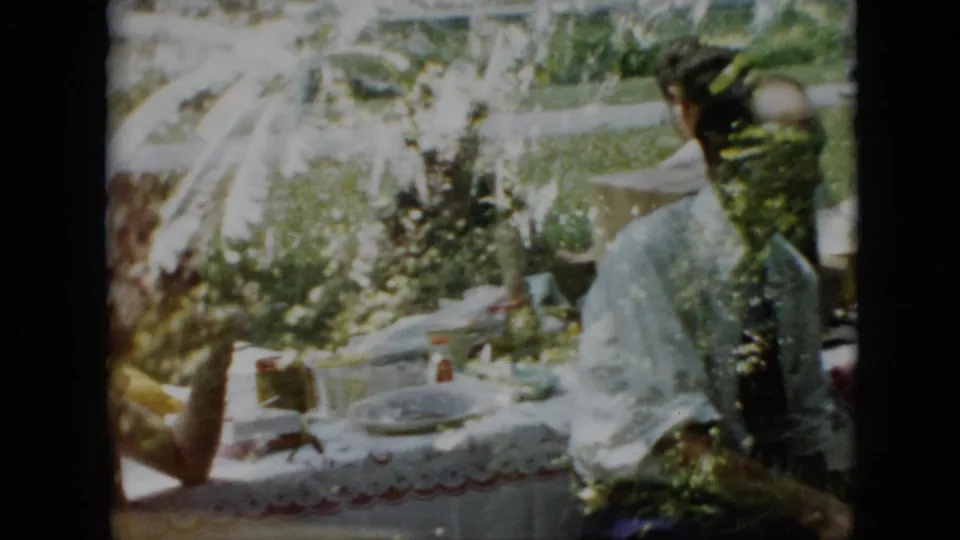 Pennsylvania 1958 home-movie frame showing a woman standing beside a family picnic table as seated adults remain gathered around plates and cups in a leafy yard.