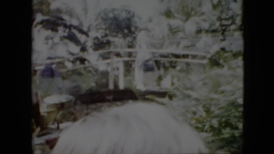 1958 Pennsylvania 8mm frame showing an adult woman in a light-colored dress carrying a purse across an arched footbridge amid trees and vegetation, with a partial foreground head visible.