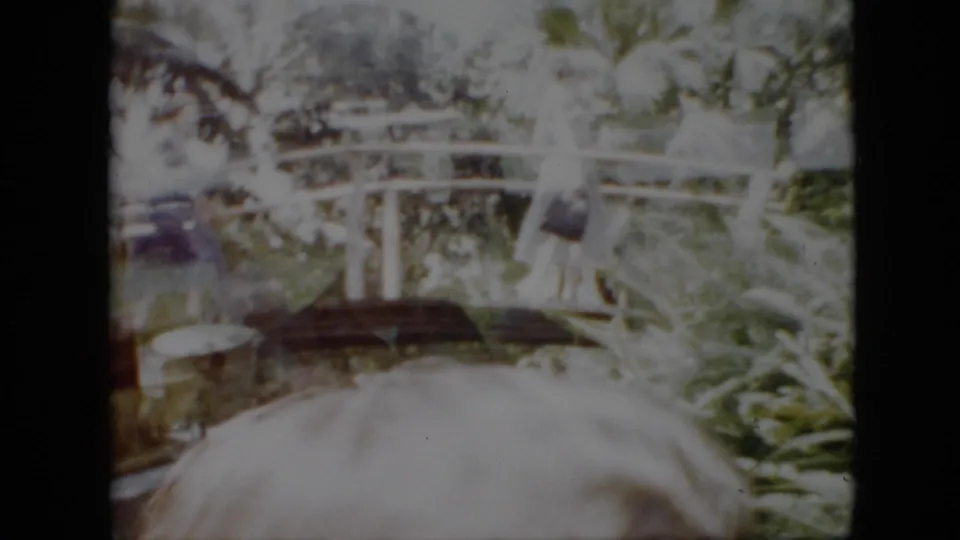 1958 Pennsylvania home-movie frame showing a white arched park bridge partly obscured by foliage and a light-haired person close in the foreground.