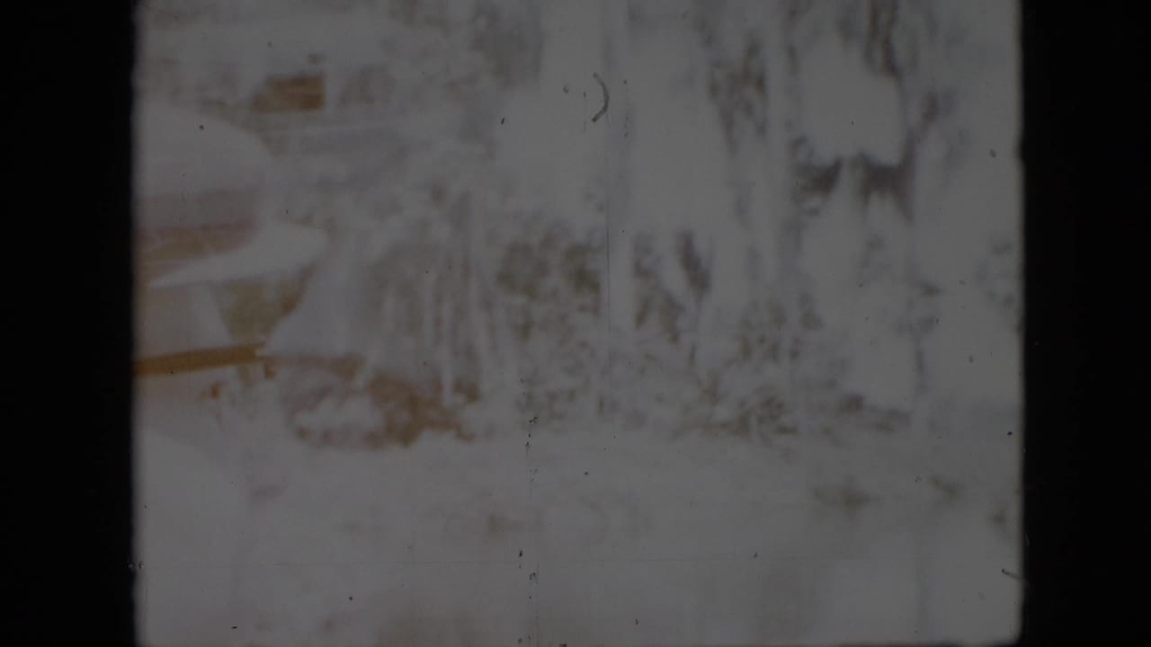 1958 Pennsylvania home-movie frame showing a pale riverbank with trees and brush viewed through a dark interior window or gate edge.