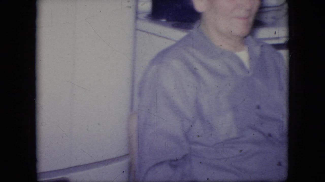 Adult man in a purple shirt seated or standing beside a white refrigerator.