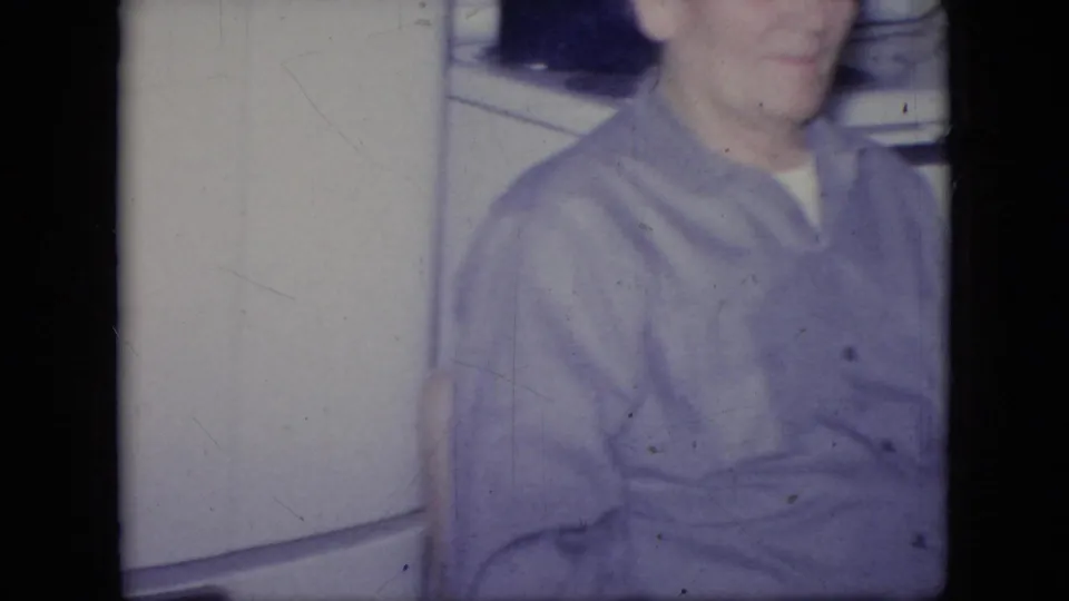 In this 1964 Philadelphia home-movie frame, an adult man in a purple button-up shirt is cropped near a white refrigerator inside a private kitchen or room.