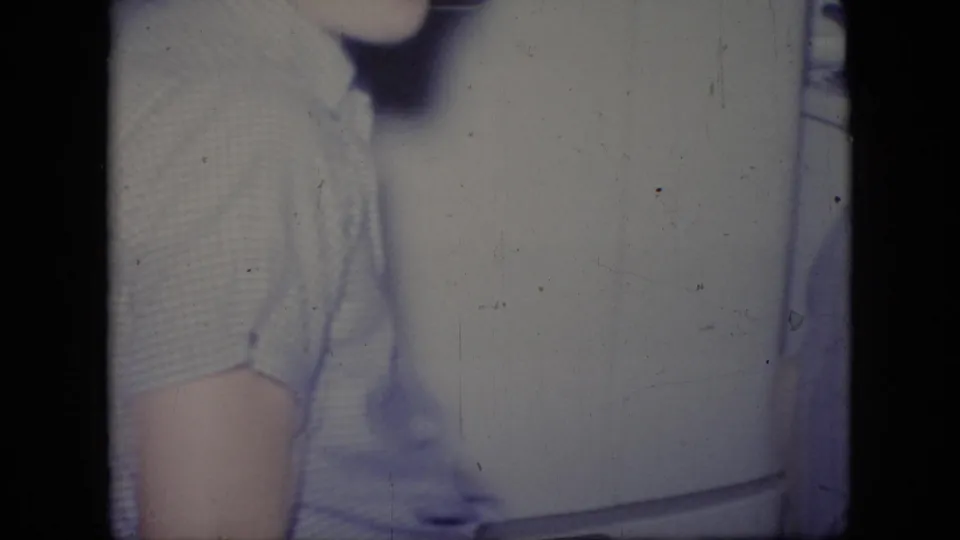 An adult man in a light striped short-sleeve shirt stands in profile near a white refrigerator in the Philadelphia home interior documented by the 1964 8mm footage.