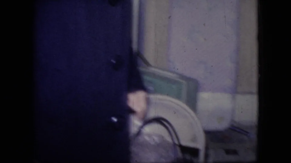 A cropped person in a dark coat or garment is partially visible near a chair or household object in the same private Philadelphia interior from the 1964 home movie.