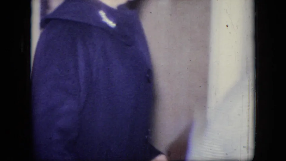 A cropped adult figure in a dark coat or heavy garment stands close to an interior wall, with the home-movie frame cutting off the face and lower body.