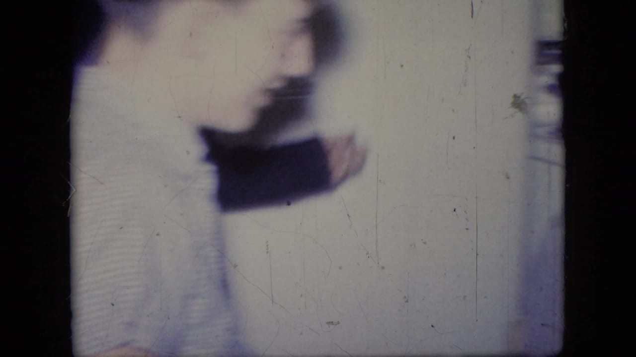 A blurred adult woman, likely in a light skirt and dark sweater, moves across an indoor Philadelphia home-movie scene from 1964.