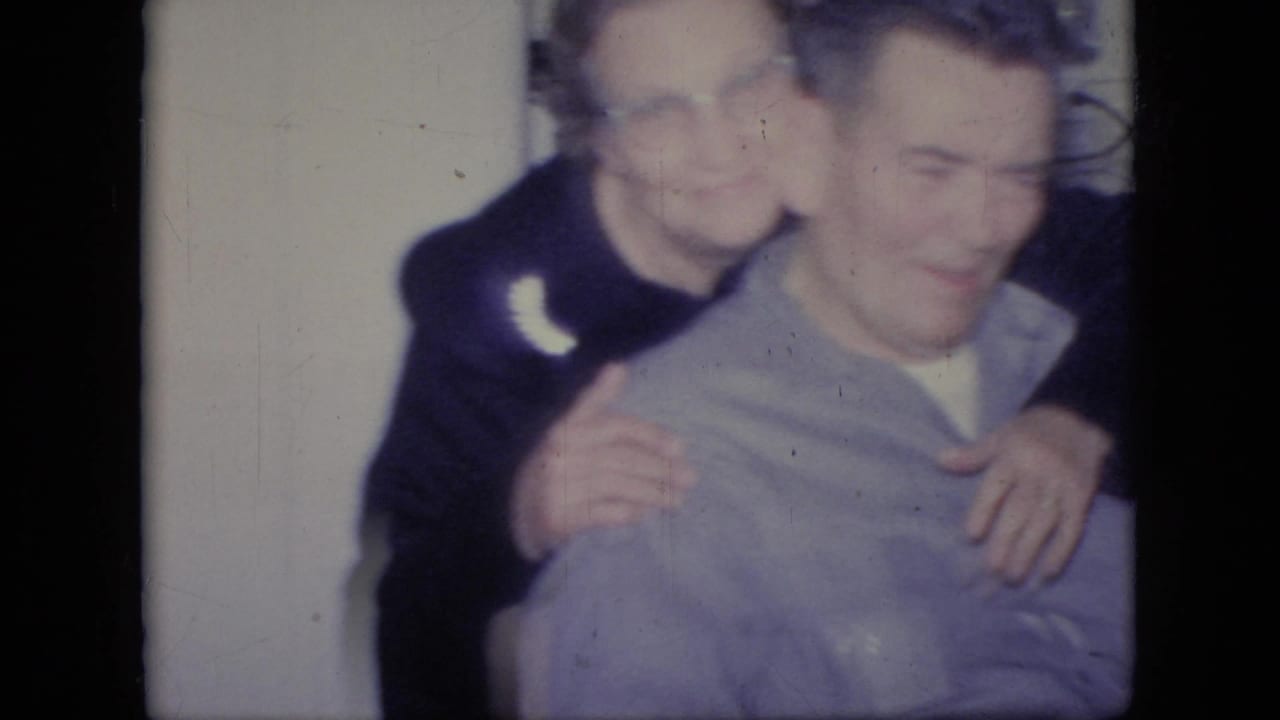 Two smiling adults embrace near a doorway or wall in a 1964 Philadelphia 8mm home-movie family gathering.