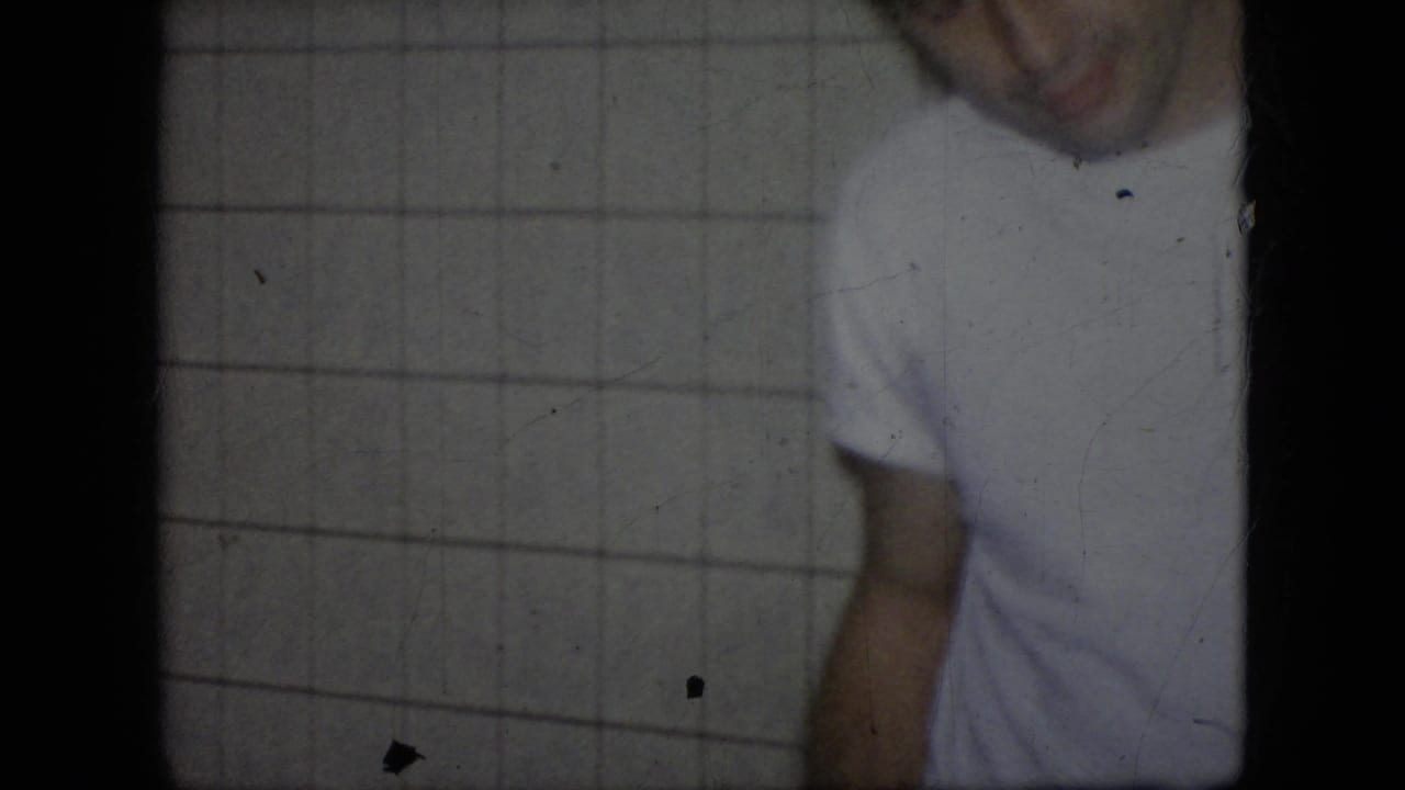 In this 1964 Philadelphia home-movie frame, an adult man in a white T-shirt and dark pants appears cropped and soft-focused against a tiled interior wall.