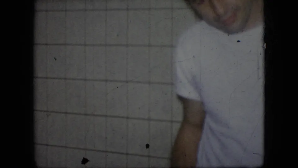 In this 1964 Philadelphia home-movie frame, an adult man in a white T-shirt and dark pants appears cropped and soft-focused against a tiled interior wall.