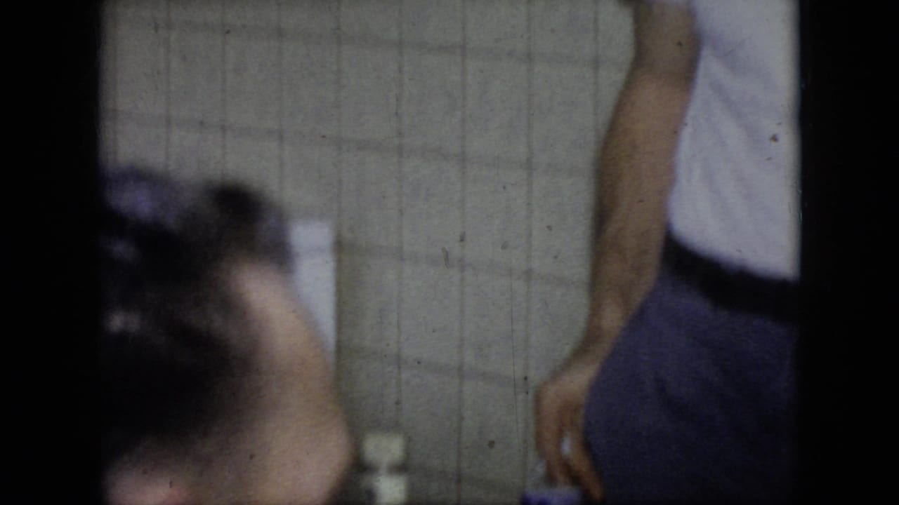 A soft-focused 1964 Philadelphia frame shows a man leaning forward near another cropped adult figure, with tiled wall surfaces and a small container in the background.