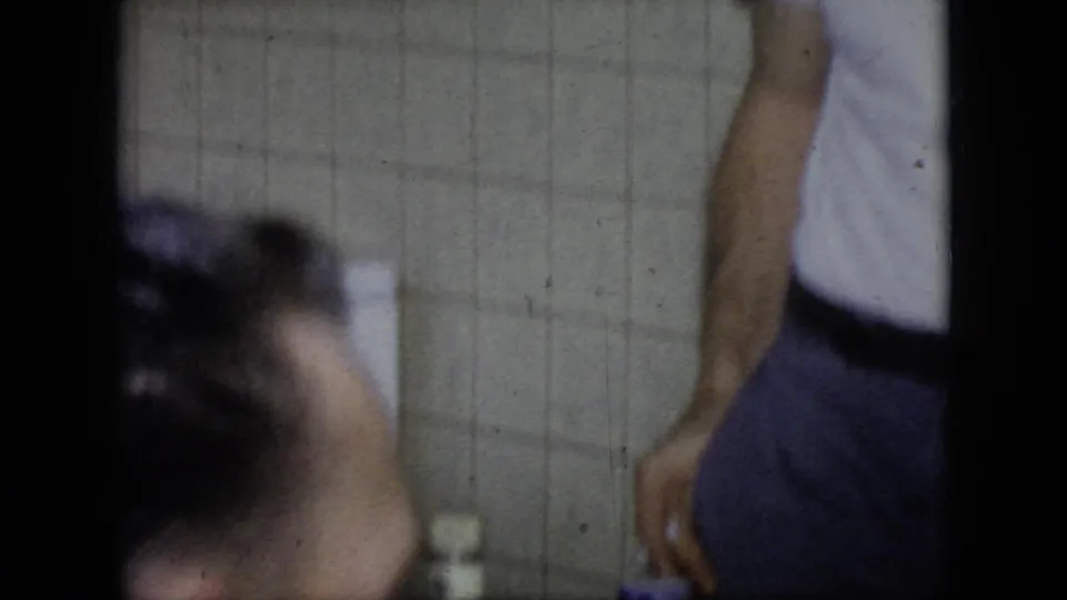 A soft-focused 1964 Philadelphia frame shows a man leaning forward near another cropped adult figure, with tiled wall surfaces and a small container in the background.