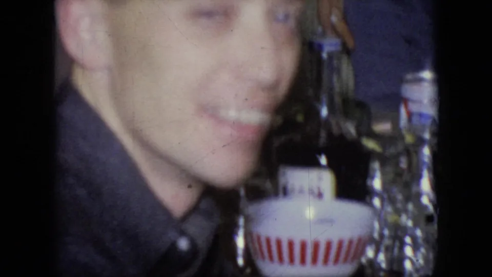 An adult man in a dark shirt smiles close to the camera in this 1964 Philadelphia home-movie frame, with glass bottles and a small container visible beside him.