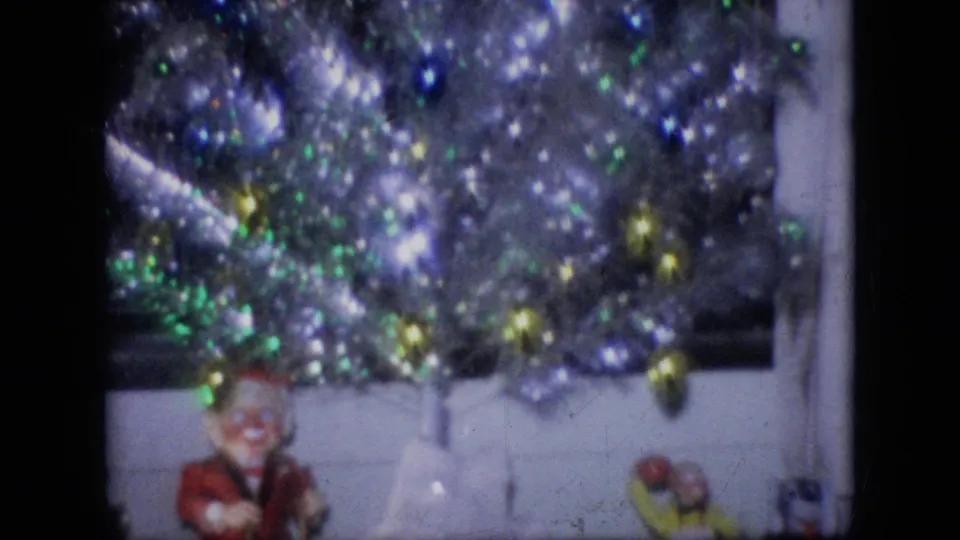 1964 Philadelphia home-movie frame showing a silver tinsel Christmas tree with colored lights, ornaments, and small holiday figurines below.