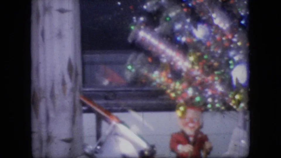 1964 Philadelphia home-movie frame showing a silver tinsel Christmas tree with colored lights near a fish tank and a small Santa-like holiday figure.