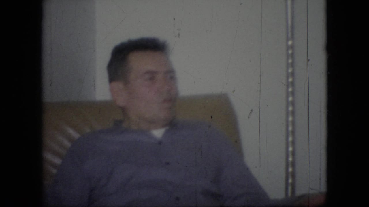 In 1964 Philadelphia home-movie footage, an adult man in a purple sweater sits in a tan upholstered armchair against a plain interior wall.