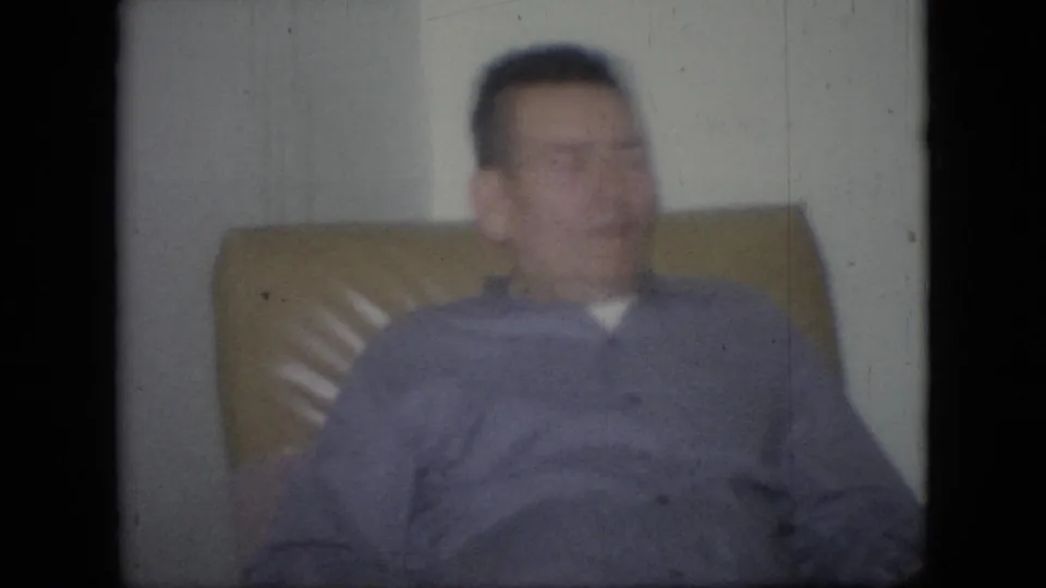 A soft 8mm frame shows the seated adult man in a purple sweater and tan chair inside a private Philadelphia home interior.
