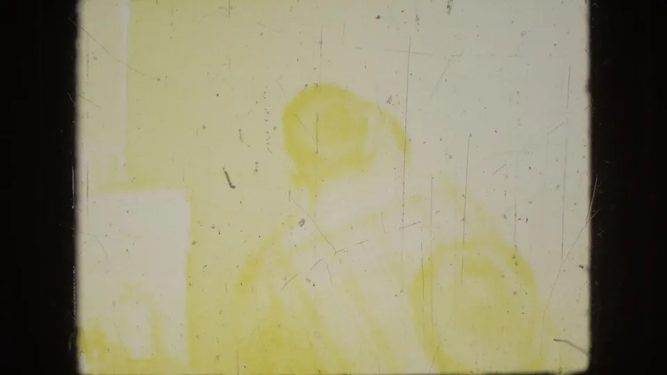 Heavily overexposed and yellow-faded 8mm frame with faint human-like forms or objects, making the domestic scene difficult to identify with confidence.