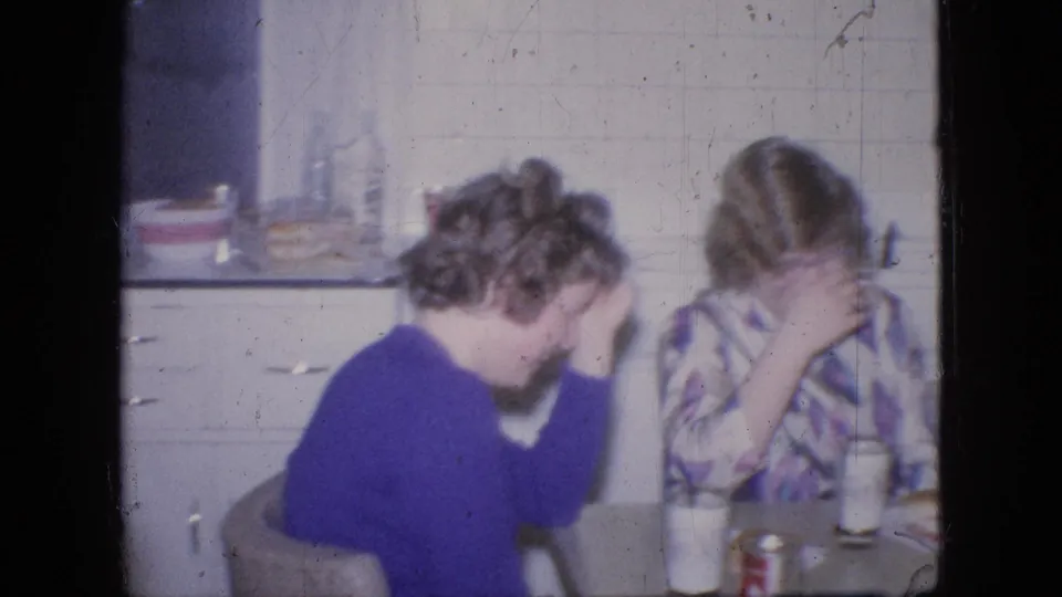 1964 Philadelphia home-movie frame showing two people seated at a kitchen table in a private interior, with one in a purple sweater and one in a patterned shirt covering their face while laughing.