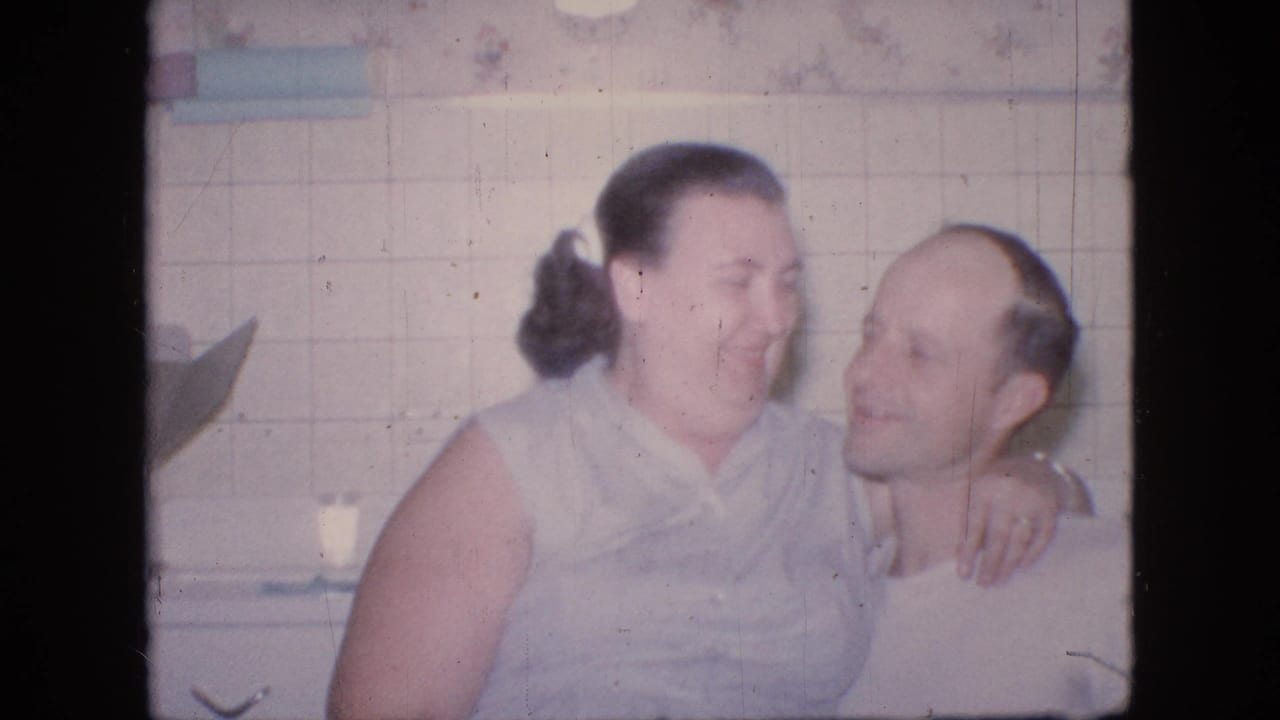 In a 1964 Philadelphia home-movie frame, a woman in a light sleeveless dress sits on a balding man's lap in a tiled kitchen while they smile and lean close.