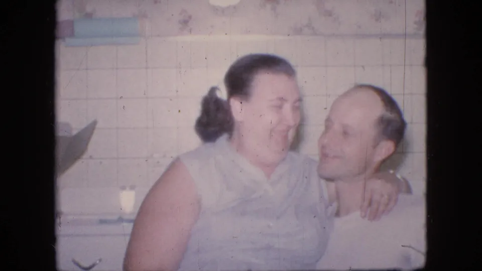 In a 1964 Philadelphia home-movie frame, a woman in a light sleeveless dress sits on a balding man's lap in a tiled kitchen while they smile and lean close.
