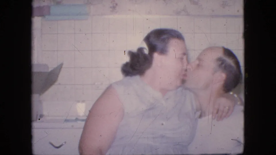 In a 1964 Philadelphia home-movie frame, the seated couple shares a kiss in a tiled kitchen, with the woman's arm around the man.