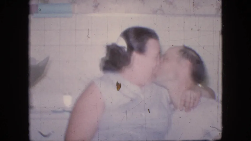 In a 1964 Philadelphia home-movie frame, the woman leans across the seated man for a kiss, with kitchen tile, drawers, and soft film blur visible around them.