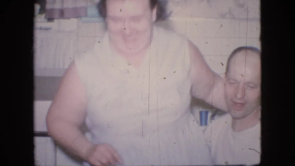 In a 1964 Philadelphia home-movie frame, the woman shifts upward beside the seated man, smiling as the tiled kitchen and countertop remain in the background.