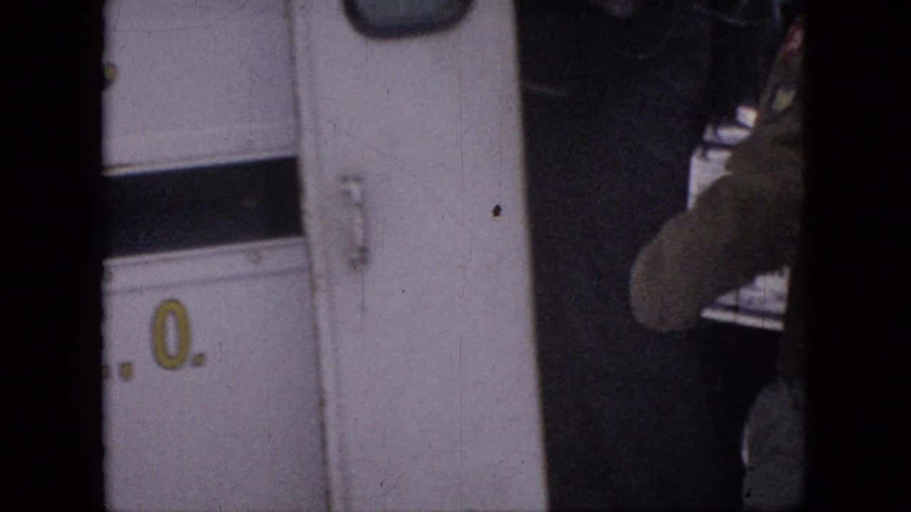 A partially visible person stands near or opens a white door in 1964 Philadelphia home-movie footage, with the frame cropped tightly and dark film borders visible.