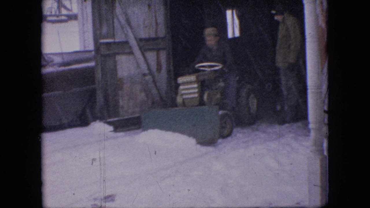 A man sits on a small tractor fitted with a green front plow blade near a shed or garage opening in a snowy Philadelphia setting.