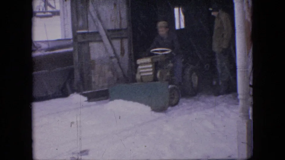 A man sits on a small tractor fitted with a green front plow blade near a shed or garage opening in a snowy Philadelphia setting.