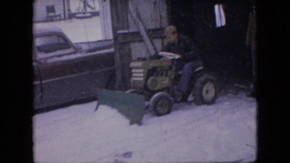 A man drives a small tractor with a green snow plow blade past a parked mid-century sedan in snow, with a shed structure behind him.