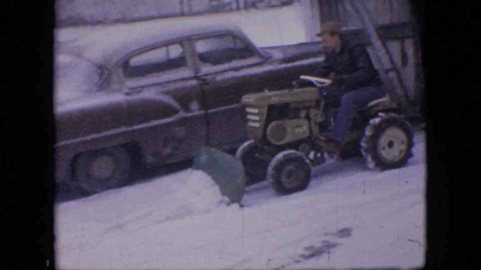 A man in a cap drives a small tractor with a green front plow through snow in front of a parked older sedan in 1964 Philadelphia home-movie footage.